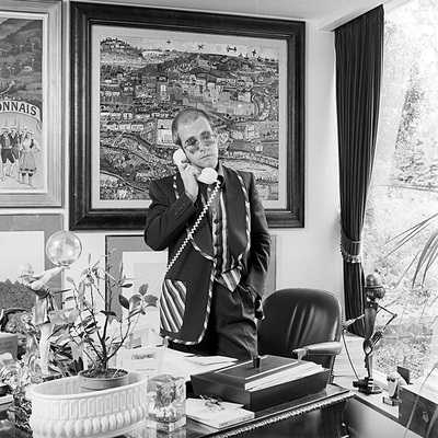 English singer and songwriter Elton John answers the phone in his study. He is wearing star-shaped sunglasses and a striped waistcoat.
